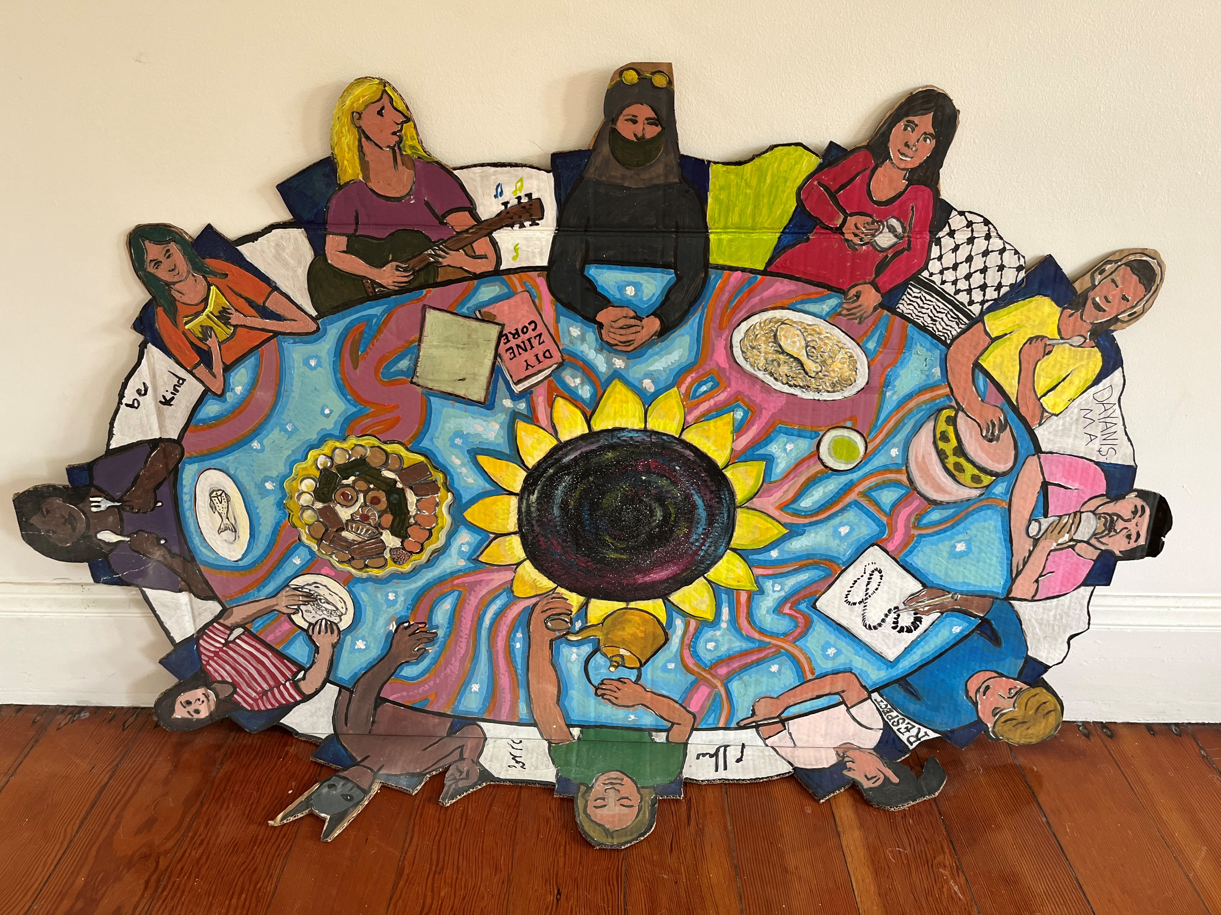 Bersama artwork, a group of people sit at a table feasting on fish, biscuits the words be kind are written in the artwork